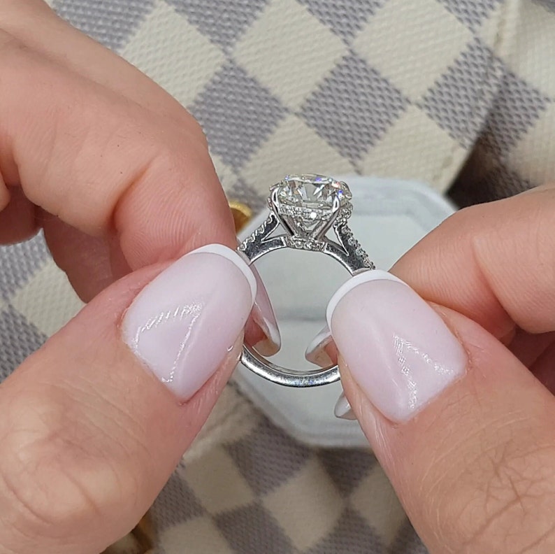 Pu&ograve; includere: Primo piano di un anello di fidanzamento in argento con un grande diamante rotondo e chiaro al centro. L'anello ha una fascia di diamanti pav&eacute; e una montatura a halo.