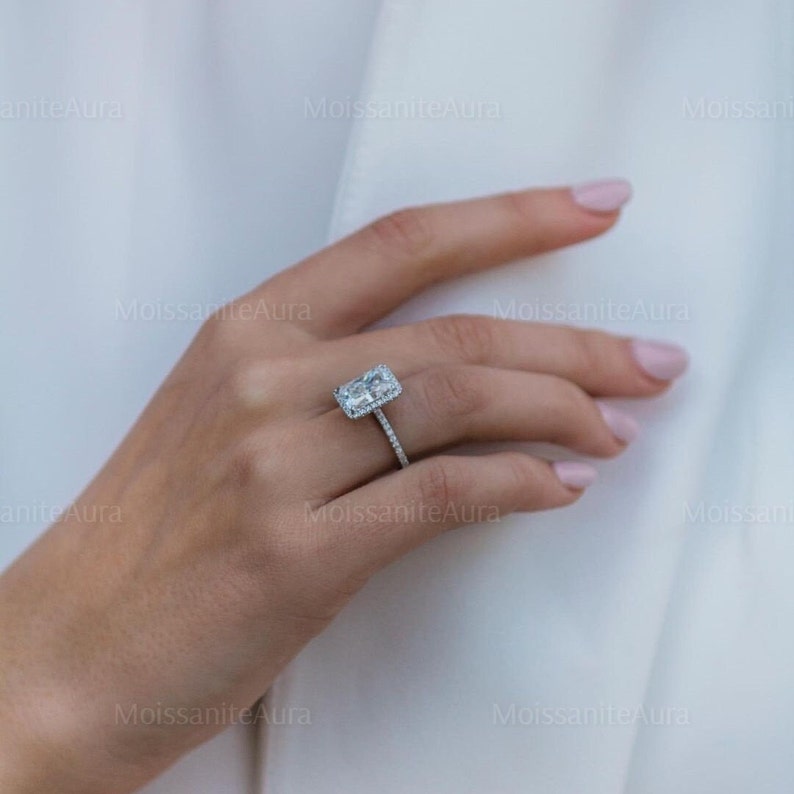 May include: A silver ring with a large, rectangular, clear gemstone surrounded by a halo of smaller gemstones. The ring is on a person's finger.