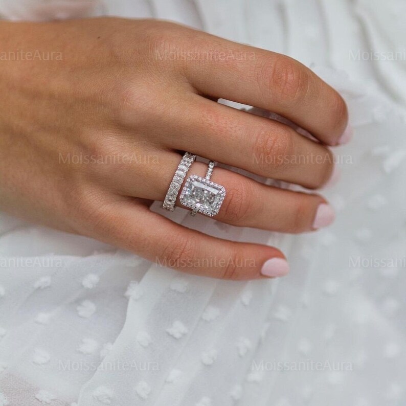 May include: A close-up of a hand wearing a silver ring with a large, rectangular, clear gemstone surrounded by smaller gemstones. The ring is worn on the ring finger of the left hand. A smaller silver band is worn on the finger next to it.