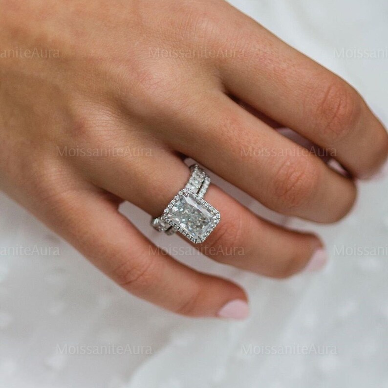 May include: A silver ring with a large, square-shaped, emerald-cut diamond. The diamond is surrounded by a halo of smaller diamonds. The ring is set on a silver band.
