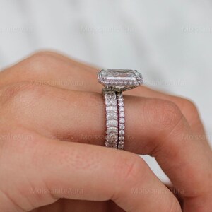 May include: A close-up of a hand wearing a silver engagement ring with a large, square-shaped diamond. The ring is set in a halo style with smaller diamonds surrounding the center stone. A matching wedding band is worn on the same finger.