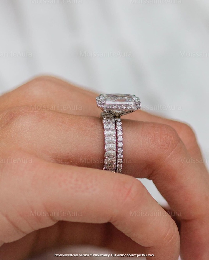 May include: A close-up of a hand wearing a silver engagement ring with a large, square-shaped diamond. The ring is set in a halo style with smaller diamonds surrounding the center stone. A matching wedding band is worn on the same finger.