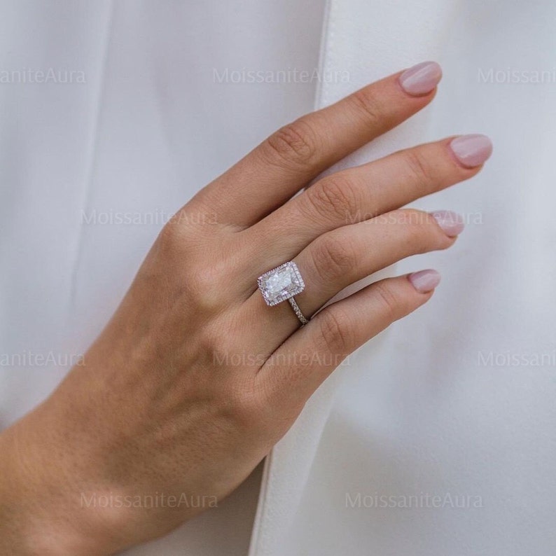 May include: A silver ring with a large, rectangular, clear gemstone surrounded by smaller, clear gemstones. The ring is on a person's finger.