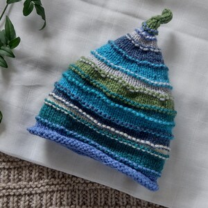 May include: A knitted hat in shades of blue, green, and white, with a small green knot at the top. The hat features horizontal stripes and a blue brim. The hat is resting on a white cloth and a textured beige surface.