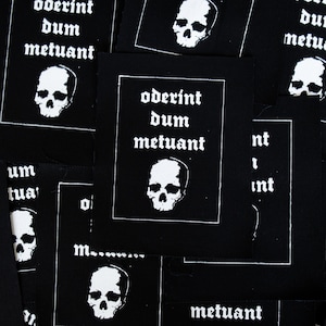 May include: A close-up of several black fabric patches with white skull and crossbones designs and the Latin phrase "oderint dum metuant" printed on them.