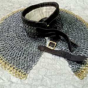 Könnte beinhalten: Ein mittelalterlicher Kettenkragen mit Leder-Halsband und Riemen. Die Kettenrüstung ist silberfarben mit einem goldfarbenen Rand. Das Leder ist dunkelbraun mit einer Messingschnalle.