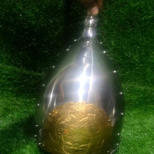 May include: A silver helmet with a brass design on the front. The helmet is on a green grassy surface.