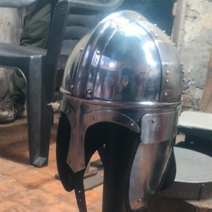 May include: A silver helmet with a pointed nose guard and a black leather strap. The helmet is on a black stand with a round base.