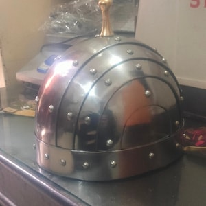 May include: A silver helmet with a gold knob on top. The helmet has a rounded shape and is decorated with small metal studs.