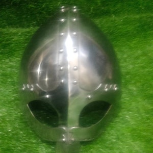 May include: A silver Viking helmet with rivets and a nose guard. The helmet is on a green background.