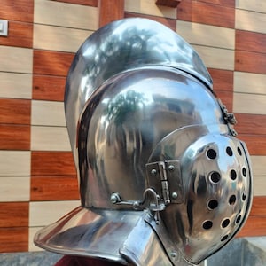 Puede incluir: Un casco medieval pulido de color plateado con una visera con múltiples agujeros pequeños. El casco tiene una parte superior redondeada y un protector de cuello acampanado. La imagen muestra la vista lateral del casco, destacando su intrincado diseño y superficie reflectante.