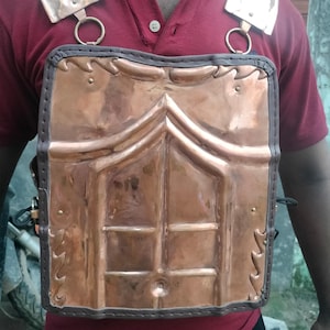 May include: A copper-colored breastplate with shoulder guards. The chest armor features a rectangular design with a series of vertical and horizontal lines, creating a grid-like pattern. The shoulder guards are rectangular with circular rings.