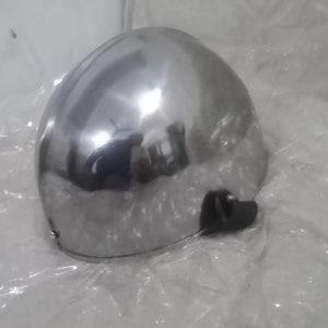 May include: A shiny silver helmet with a black strap and buckle. The helmet is made of metal and has a smooth, reflective surface.