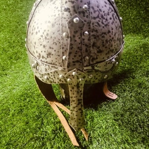 May include: A grey helmet with a textured surface and metal rivets. The helmet has a leather strap and is on a green background.