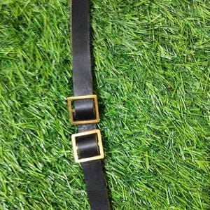 May include: A black leather strap with two gold metal buckles. The strap has a loop at each end.