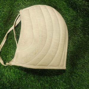 Puede incluir: Un gorro acolchado beige con detalles de costuras curvas. El gorro tiene forma redondeada y lazos a cada lado. La tela parece ser de fibra natural, sobre un fondo verde.
