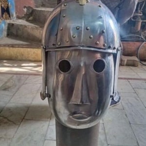 May include: A silver metal helmet with a face mask and a pointed top. The helmet has a textured surface and is decorated with small rivets.