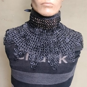 May include: A black chainmail gorget featuring interlocking metal rings, worn around the neck and shoulders. The piece is displayed on a mannequin head and torso wearing a black and gray horizontally striped shirt with visible text on the chest.