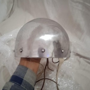 May include: A silver helmet with a rounded top and a leather strap. The helmet has several rivets around the edge.
