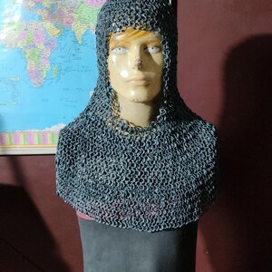 Puede incluir: Un coif de cota de malla, un tipo de armadura medieval para la cabeza y el cuello, se exhibe en un maniquí. El coif está hecho de pequeños anillos de metal entrelazados y cubre la cabeza, el cuello y los hombros. El maniquí está frente a un mapa del mundo.