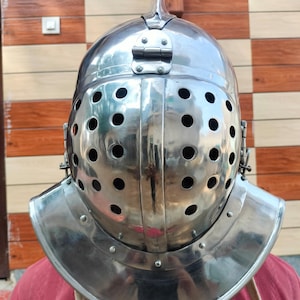 Puede incluir: Un casco de metal plateado pulido con una parte superior puntiaguda y numerosos agujeros circulares para los ojos. El casco tiene una protección para el cuello y es probablemente una pieza histórica o de disfraz. El fondo es una pared de madera.