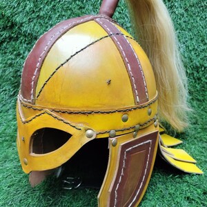 May include: A yellow leather helmet with brown leather accents and a long blonde hairpiece. The helmet has a face mask and ear flaps.