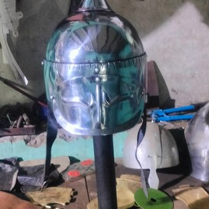 May include: A silver helmet with a pointed top and a decorative design on the front. The helmet is on a stand and there are other leather and metal objects in the background.