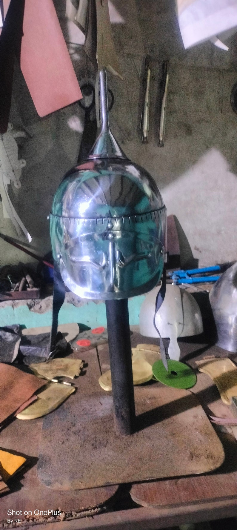 May include: A silver helmet with a pointed top and a decorative design on the front. The helmet is on a stand and there are other leather and metal objects in the background.