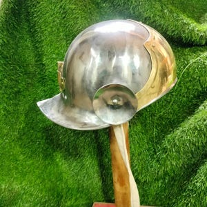 Puede incluir: Un casco plateado pulido con detalles de latón, con visera y protección para los oídos. El casco se exhibe sobre un soporte de madera con un detalle de tela blanca, sobre un fondo verde.