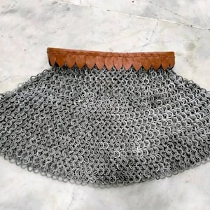 Può includere: Un pezzo di armatura a maglia di catena con un colletto in pelle marrone. La maglia di catena è composta da piccoli anelli di metallo ad incastro. Il colletto è marrone scuro con un bordo smerlato. La maglia di catena è di colore argento.