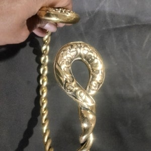 Puede incluir: Un brazalete de metal retorcido de color dorado con un cierre decorativo y adornado. El cierre tiene forma de bucle con un diseño floral.