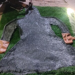 May include: A long-sleeved chainmail hauberk constructed from interlocking metallic rings, featuring a hood and integrated brown leather mittens at the ends of the sleeves. The garment is laid flat, displaying a flared skirt and a full-body design.