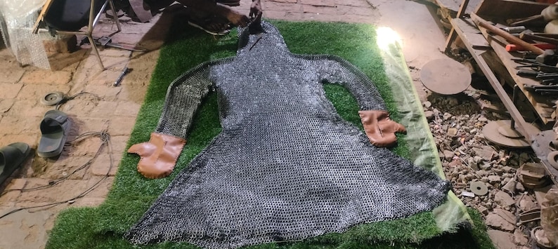 May include: A long-sleeved chainmail hauberk constructed from interlocking metallic rings, featuring a hood and integrated brown leather mittens at the ends of the sleeves. The garment is laid flat, displaying a flared skirt and a full-body design.