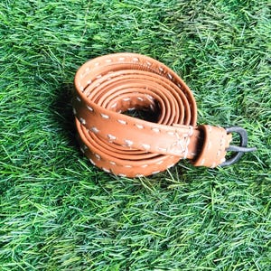 Puede incluir: Un cinturón de cuero marrón claro enrollado, con costuras blancas y una hebilla de metal negro. El cinturón está sobre césped artificial verde. Un accesorio clásico.