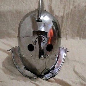 Puede incluir: Un casco plateado con una cresta puntiaguda y dos agujeros para los ojos. El casco tiene una correa para la barbilla y está hecho de metal.
