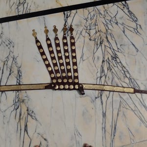 May include: A brown leather belt with multiple straps, each adorned with gold-colored circular accents. The belt features a gold-colored buckle and decorative elements. The design suggests a historical or costume-related item.