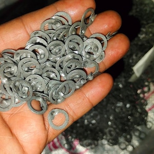May include: A handful of small, gray, metal washers. The washers are circular with a split on one side. The image shows a close-up view of the washers, with some in focus and others blurred in the background.