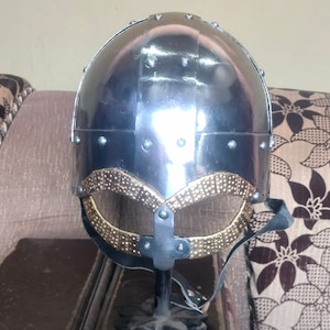 May include: A silver and gold Viking helmet with a black leather strap. The helmet is on a black stand and has a decorative design around the top.