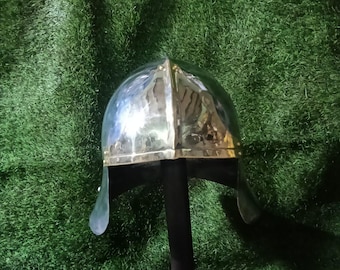 May include: A polished metal helmet with a rounded top and a brass-colored band around the base. The helmet has cheek guards and is displayed on a black stand against a green background.