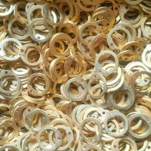 May include: A close-up of a large quantity of gold-coloured metal rings, likely washers or spacers, in a black plastic bag. The rings are stacked and overlapping, creating a dense, textured pattern.