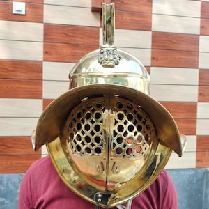 Könnte beinhalten: Ein polierter Gladiatorhelm aus Messing mit einem markanten Gesichtsschutz mit einer Reihe kreisförmiger Perforationen. Der Helm hat einen Kamm und ein dekoratives Emblem oben. Das Bild zeigt den Helm von vorne, mit einer Person, die ein rotes Hemd trägt.