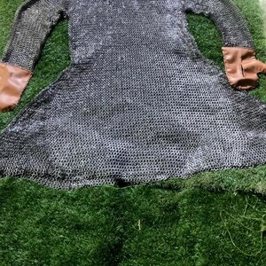 May include: A long-sleeved chainmail hauberk constructed from interlocking silver-toned metal rings, featuring an integrated hood and tan leather cuffs at the end of each sleeve. The garment is laid flat, displaying a flared skirt and a neck opening.