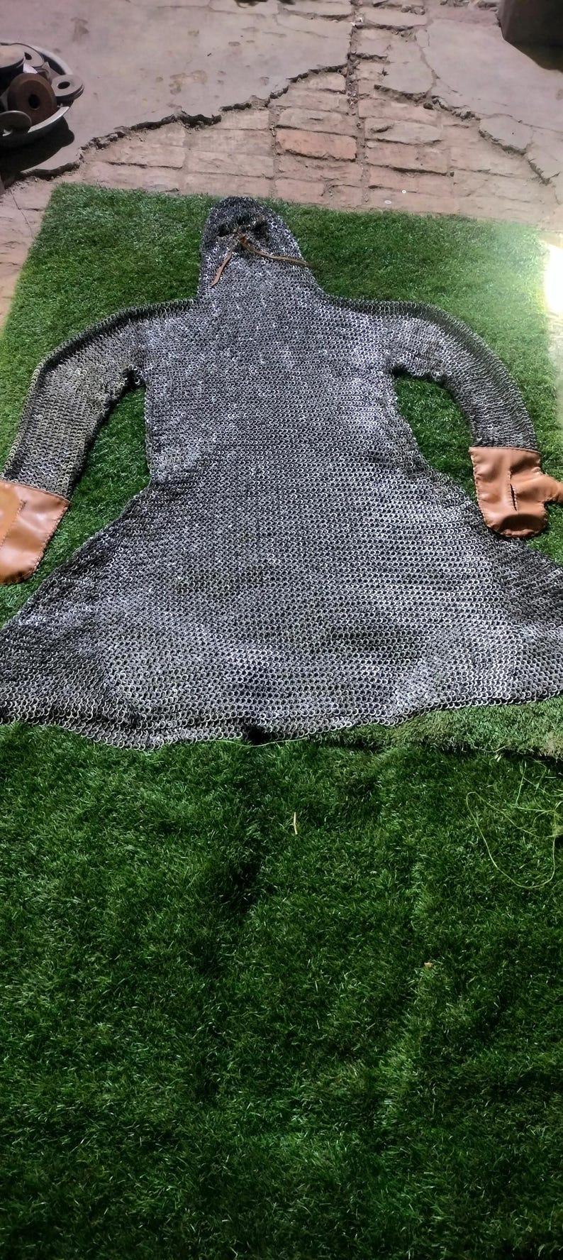 May include: A long-sleeved chainmail hauberk constructed from interlocking silver-toned metal rings, featuring an integrated hood and tan leather cuffs at the end of each sleeve. The garment is laid flat, displaying a flared skirt and a neck opening.