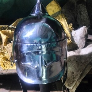 May include: A silver helmet with a pointed top and a face mask. The helmet is on a wooden stand.