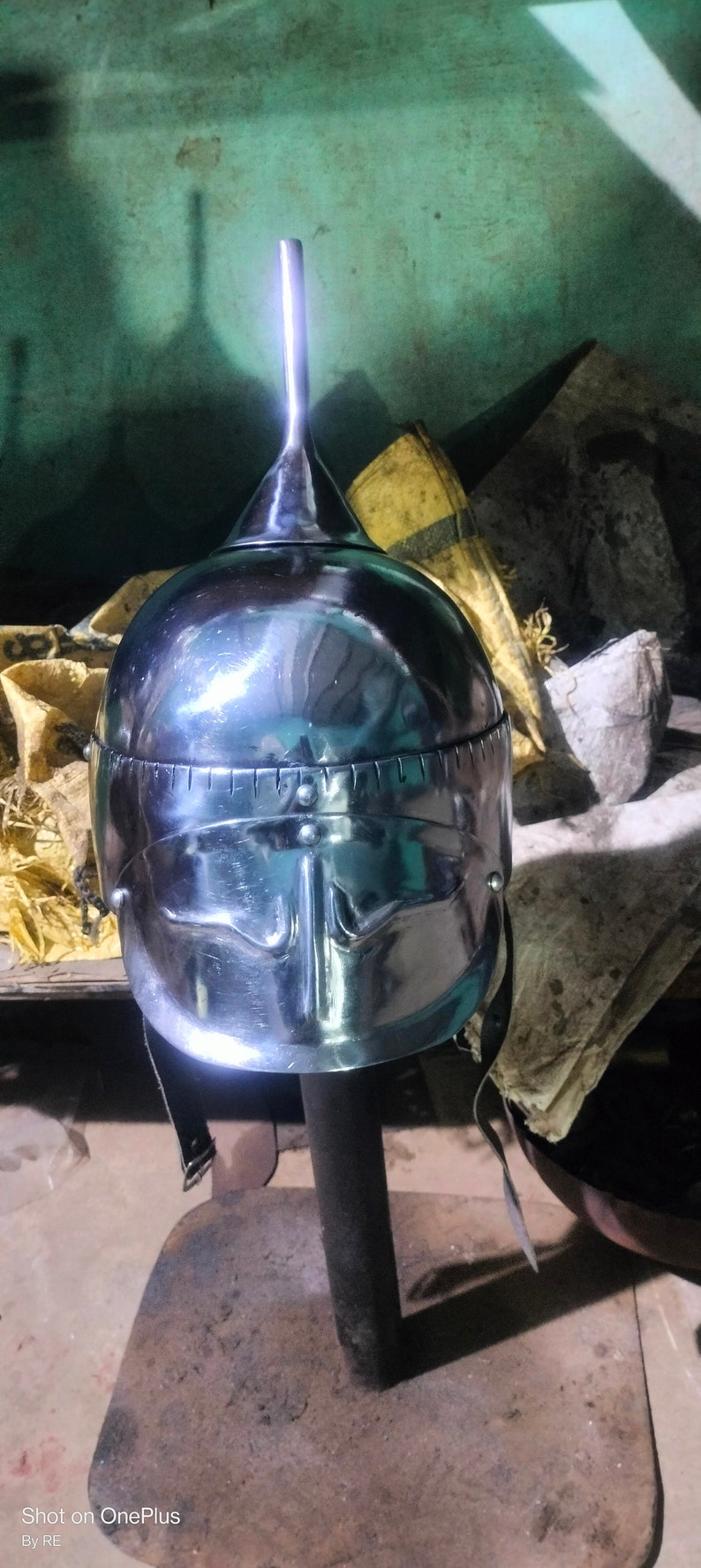 May include: A silver helmet with a pointed top and a face mask. The helmet is on a wooden stand.