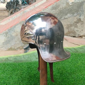 May include: A polished silver-coloured metal helmet with a smooth, rounded dome and a flared neck guard. The helmet features decorative embossed side panels secured with small rivets and includes a brown leather chin strap hanging from the interior.