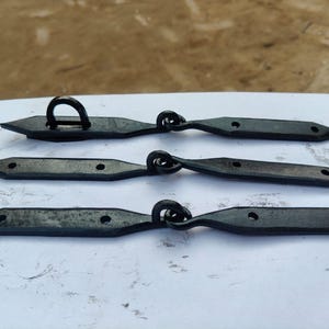 Pode incluir: Três dobradiças de porta de metal preto com uma extremidade pontiaguda e um laço de um lado. As dobradiças estão dispostas em uma fila sobre uma superfície branca.