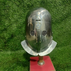 Puede incluir: Un casco medieval pulido de color plateado, con una parte superior redondeada y una visera con dos orificios circulares para los ojos. El casco está montado sobre un soporte de madera con una base roja, sobre un fondo verde.