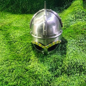May include: A silver helmet with a pointed top and a decorative band around the bottom. The helmet has a visor with a yellow and black pattern.