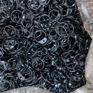 May include: A close-up image of a large quantity of black metal rings. The rings are all the same size and shape, and they are piled up in a plastic bag.